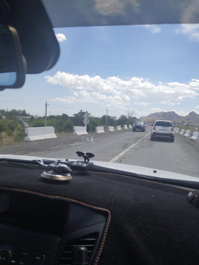 The image, captured from inside a vehicle, shows a view of a paved road in Asmansay, Uzbekistan. The road has some cracks and appears to be a two-lane highway with white concrete barriers lining the side. A white SUV with the license plate 95KX202NA is in the right lane, followed by a dark-colored car in the left lane. Further down the road, more cars are visible. The environment is dry and somewhat barren, with scattered trees and bushes on the left side of the road. In the distance, mountains are visible under a partly cloudy blue sky. The dashboard of the vehicle is in the foreground, with a miniature silver helicopter and a black electronic device mounted on it.