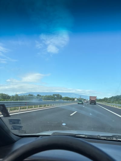 The image shows a view from the driver's seat of a car traveling on a highway. The sky is blue with scattered clouds. In the distance, there are mountains and trees. The highway has multiple lanes, and other vehicles are visible, including cars and a truck. The scene appears to be in or near Dolni Bogrov, Bulgaria, as indicated by the highway leading towards the area.