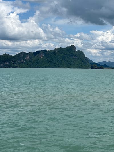 The image shows a vast expanse of greenish-blue water in the foreground, with gentle waves rippling across its surface. In the midground, a lush green, mountainous island rises dramatically from the sea, its slopes covered in dense vegetation and rocky outcrops. The sky above is a mix of dramatic, heavy grey clouds and patches of bright blue sky, suggesting a dynamic weather system. The scene is likely located off the coast of Thong Nian, Thailand, a region known for its picturesque islands and clear waters. No people or specific objects are visible in the frame, focusing solely on the natural landscape.