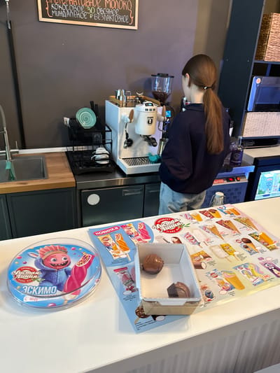 A person with long brown hair pulled into a ponytail, wearing a dark blue top and light blue jeans, stands behind a coffee machine in a dimly lit cafe. The person is seen from the side, looking down and to the left. The cafe appears to be modern, with dark gray walls and wooden accents. On the counter are various items, including a coffee grinder, a white coffee machine with a portafilter, and a black metal rack holding plates and a green saucer.
To the right of the coffee machine, there is a display of ice cream products. These include several ice cream bars and cones with colorful packaging. A small cardboard box displays two chocolate-covered ice cream treats, one of which has been partially eaten. A circular container of "Эскимо" (Eskimo) ice cream, featuring a cartoon character, is prominently placed in the foreground. Some visible text on the packaging is in Russian, mentioning "Чистая Линия" (Chistaya Liniya - Clean Line) as a brand, and "Эскимо" (Eskimo). Other text indicates different ice cream flavors, such as "кокосовое" (coconut), "миндальное" (almond), and "безлактозное" (lactose-free). There is also a chalkboard sign on the left wall with handwritten text in Russian, listing milk, coconut, almond, and lactose-free options. The overall scene suggests a typical day in a cafe or ice cream shop.