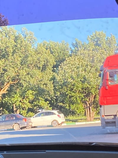 The media file shows a street scene in Yekaterinovka, Russia, likely during daylight. The image is oriented vertically, suggesting it was captured from a slightly elevated perspective or the device was held sideways.
In the foreground, a bright red truck is partially visible at the top. Below the truck, a dense canopy of green trees dominates the left side of the frame, indicating a leafy, possibly summer or early autumn setting. The sky above is a clear, light blue with some subtle imperfections.
To the right of the trees, a paved road is visible. Parked on the road are two cars. The upper car is a light-colored hatchback, possibly white or beige, with its rear end facing towards the viewer. Below it, a darker colored car, possibly dark blue or black, is also parked. Both cars have their wheels visible, and their general shapes suggest modern passenger vehicles.
The immediate area around the cars appears to be a roadside. There is a thin strip of green and white border between the road and what seems to be a grassy verge or sidewalk. In the middle ground, behind the parked cars and to the right of the trees, there is a pile of logs or thick branches, suggesting recent activity such as logging or landscaping.
No people are clearly visible in the image. The overall impression is a quiet street with parked cars adjacent to a verdant natural area. The lighting suggests it's daytime, and the clear sky hints at fair weather. No text is discernible in the image.