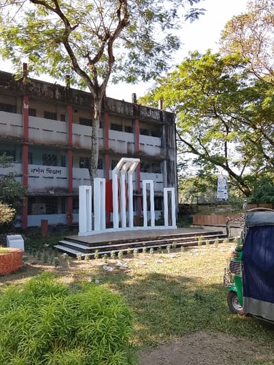 The image captures an outdoor scene in what appears to be an educational or public campus in Shibpur, Bangladesh. A white Shaheed Minar (Martyr's Monument), featuring a central red disc flanked by several white vertical pillars of varying heights, stands on a dark-edged, stepped platform in the mid-ground. In the background, a three-story, light-colored concrete building with red vertical support beams shows signs of age and wear. The Bengali text "দর্শন বিভাগ" (Dorshon Bibhag, meaning Philosophy Department) is visible on the building's facade. A large tree with a prominent, gnarled trunk and leafy canopy rises in front of the building, partially obscuring it and the monument. The foreground consists of sparse grass and dirt patches, with a dense green shrub to the lower left. Scattered white debris is visible on the ground near the monument's base. On the far right, a green and blue auto-rickshaw is partially visible. Lush green foliage from other trees is present on the right side and in the distant background. The scene is illuminated by bright daylight, casting shadows to the left, indicative of morning or late afternoon. No individuals are visible.