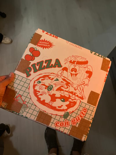 The image shows a pizza box held in a person's hand. The box is printed with a cartoon image of a chef and a pizza. The words "PIZZA" and "con Gusto" are also printed on the box. The box appears to be sealed with brown paper tape. Parts of two pairs of legs, one with white shoes and another with black shoes, and some dark spots are visible in the bottom part of the frame. The location in the city of San Pedro Cholula, Mexico cannot be determined from the image.