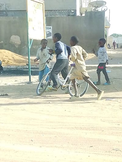 Four young boys are on a dirt road near a building with a sign. One boy is on a white bicycle, while the other three stand nearby. The boy on the bicycle is wearing gray pants and a blue shirt. One of the other boys is wearing a light shirt and blue jeans, while the other two boys are wearing long-sleeved shirts. One boy is walking with a pair of sandals. Several other people are visible in the background.