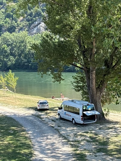 The image depicts a serene outdoor scene next to a body of water. In the foreground, a large tree with lush green foliage casts dappled shadows on the ground. Parked nearby is a white camper van with its rear door open, suggesting it might be in use or being prepared for a trip. A second, smaller white car is parked behind the van.

In the middle ground, several people are visible near the water's edge, some appearing to be relaxing or wading in the shallows. The water itself is a calm, green hue, reflecting the surrounding environment. The background reveals a lush, tree-covered hillside that rises steeply from the water, with hints of rocky outcrops.

The lighting suggests it is a sunny day, possibly late morning or afternoon, given the long shadows. The overall atmosphere is tranquil and recreational, hinting at a day spent enjoying nature by the water.

Visible text on the side of the camper van appears to be a logo with some lettering, but it is not entirely legible. The license plate on the smaller car is also too blurry to read. The location is Zvornik, Bosnia and Herzegovina, which aligns with the mountainous terrain and river setting.