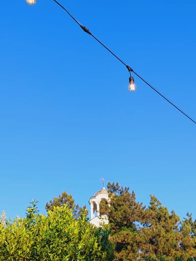 The image shows a clear blue sky with a string of lights extending diagonally from the top left to the middle right. Below the lights, a white bell tower with a cross on top is visible, partially obscured by lush green trees. The bell tower appears to be part of a religious building, possibly a church, and its architecture suggests a European style. The trees in the foreground are dense and vibrant green, creating a natural contrast with the man-made structures and the clear sky. The overall scene is peaceful and evokes an outdoor setting, possibly in a park or a residential area. The location is Grivitsa, Bulgaria, as indicated.