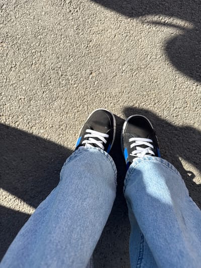 A downward-angle shot shows a pair of feet clad in light blue denim jeans and black suede sneakers with white laces and a blue stripe. The shoes are positioned on a concrete surface. Shadow patterns partially obscure both the ground and the denim. The image contains no indicators that would reveal its location in Krilovo, Russia.