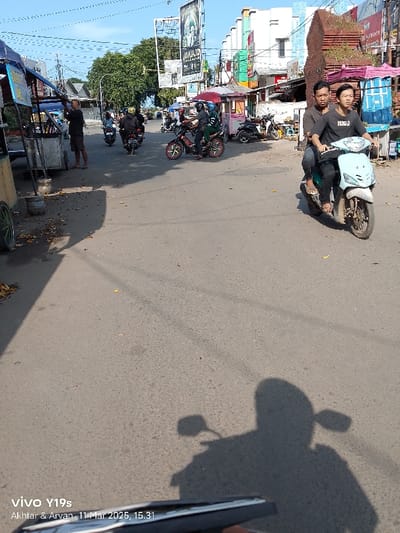 The image, taken in Cirebon, Indonesia (latitude: -6.725, longitude: 108.571), shows a street scene with several people on motorcycles. Two men, both wearing black shirts, are on a blue motorcycle, traveling to the right. There are other motorcycles in the distance, some stationary and some moving. The street is asphalt and there are buildings on both sides. The image is captured from the perspective of a person on a motorcycle, looking towards the street. The shadow of the motorcycle the image was taken on can be seen in the foreground. There are power lines overhead and some trees are visible in the distance. The image was taken during the day and the sky is clear.