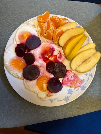 A plate of food is arranged on a light gray, textured surface, likely a tablecloth or placemat. The plate appears to be white with a blue floral pattern around the edge. The food items include: sliced apples (yellowish color), mandarin orange segments, fried eggs (several), sliced beets (dark purple), and a piece of cooked meat or other protein (pinkish-red). The eggs have been topped with the sliced beets and the pinkish-red meat. The scene does not provide sufficient information to verify the location as Oswestry, United Kingdom.