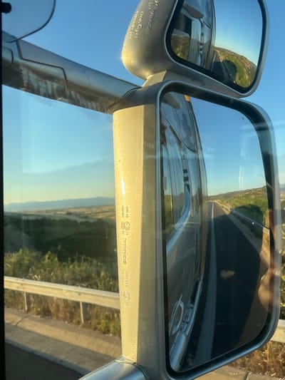 The image depicts a view from inside a vehicle, looking outward and into side mirrors. The scene is set in Avra, Greece, on a clear, sunny day with a bright blue sky. The foreground shows a portion of the vehicle's interior, including a metallic bar and textured vertical support.
In the side mirrors, a highway is visible, curving through a hilly, green landscape with patches of dry vegetation. The road appears to be asphalt with a white dividing line. The vegetation suggests a rural or semi-rural setting, consistent with the outskirts of a town or along a main transit route.
One of the mirrors reflects the vehicle's interior and the passing scenery, including the road ahead and the surrounding hills. Markings are visible on the vehicle's support structure, including what appears to be "SCANIA" and some alphanumeric codes, suggesting the vehicle is a Scania truck or bus. There is no indication of people or any interaction occurring. The environment is natural, with the road and vegetation being the dominant elements outside the vehicle.