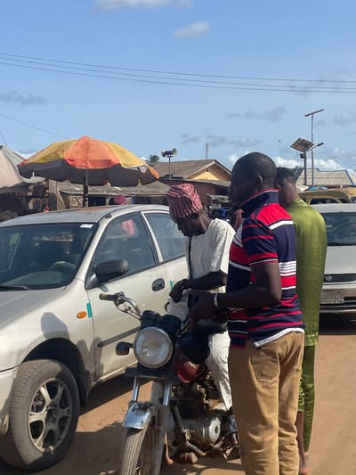 The image shows three men near a white car and a motorcycle in Ajibo, Nigeria. One man, dressed in a red and blue striped shirt and khaki pants, appears to be working on the motorcycle. Another man, in a white shirt and red patterned head covering, is also focused on the motorcycle. A third man, wearing a green shirt and khaki pants, stands behind them. A multi-colored umbrella is visible nearby, along with utility poles and power lines against a blue sky. Other vehicles and buildings are visible in the background.