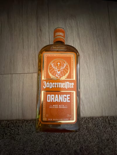 This is a photograph of a bottle of Jägermeister Orange, a flavored herbal liqueur. The bottle is lying on its side on a wooden surface, with a dark gray, fuzzy texture, possibly a rug or carpet, visible to the right of the bottle.
The bottle itself has a distinctive amber color, indicating the liquid inside. The main label is orange and white, featuring the iconic Jägermeister stag logo in the center. Bold white text on the label reads "Jägermeister" and "ORANGE". Below "ORANGE" it says "-MADE WITH - SICILIAN ORANGES". To the right of this, "33% vol" and "DER KRÄUTERLIKÖR" are visible. There is also text in German running vertically along the left side of the label, which appears to be part of the brand's slogan. The bottle cap is also orange with white lettering that reads "ORANGE".
The lighting in the image is somewhat dim, suggesting it might be indoors with artificial light or taken during the evening. The wooden floor has a visible grain and appears to be a light to medium brown color. The overall presentation is simple, focusing on the product itself. No people are visible in the image.