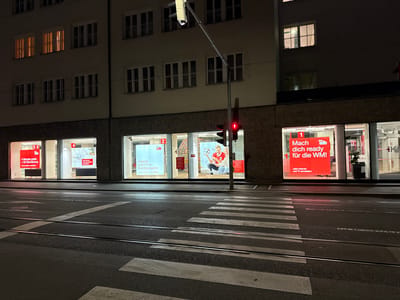 The image captures a nighttime street scene in Innsbruck, Austria. A pedestrian crossing with white stripes is prominent in the foreground, with tram tracks running parallel to it. In the background, a building with multiple stories is illuminated by interior lights shining through its windows. The ground floor of the building features several shop windows displaying advertisements. One prominent advertisement in red text reads "Mach dich ready für die WM!" (Get ready for the World Cup!). Another advertisement displays "4 Monate gratis + 5€ Strombonus" (4 months free + 5€ electricity bonus). A traffic light with a red signal is visible, casting a red glow on the wet asphalt of the street. The overall atmosphere is dark and urban, with artificial lights from the building and streetlights providing illumination.