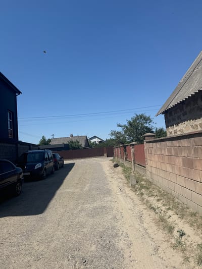 A sun-drenched, clear blue sky dominates the upper portion of the image, with a single bird in flight visible in the far left. Below, a gravel road stretches into the distance, flanked by a red metal fence and a substantial brick wall on the right. On the left side of the road, several parked cars are visible, including a dark colored van with a license plate that appears to read "3964HI". A dark building with a corrugated metal exterior is situated to the left of the cars. In the background, houses with tiled roofs and trees suggest a residential or semi-rural setting. The overall scene depicts a quiet, daytime view of a street or driveway in what appears to be a suburban or rural area, possibly in Sarny, Ukraine, given the context provided.