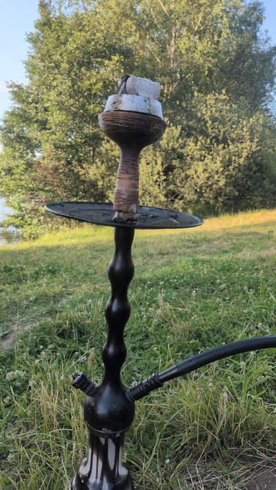 An outdoor, close-up shot shows a black hookah with a black hose attached. The hookah is sitting on green grass, with a body of water and trees in the background. The image is taken during the daytime.