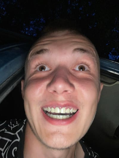 The media file is a close-up, sideways selfie of a man with light-colored skin and short, light brown hair. He is smiling broadly, showing his teeth. His eyes are open and looking towards the camera. The background is dark, with some blurry blue shapes that appear to be foliage or lights. The man is wearing a black t-shirt with a white, abstract pattern. The lighting suggests it might be nighttime or a dimly lit indoor setting. There is no visible text in the image.