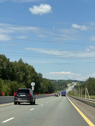 A dark-colored Volvo SUV is seen from the rear on a multi-lane highway, traveling in the right lane. The SUV has a license plate with Cyrillic characters and numbers: P 528 XY 18. The highway is flanked by a concrete barrier on the left and a guardrail on the right. Beyond the barrier, a dense line of green trees forms a forest. On the right side of the highway, utility poles with wires are visible. The sky is blue with scattered white clouds and thin power lines stretch across the upper portion of the image. Further down the highway, other vehicles, including a blue truck and several cars, are visible. The landscape features rolling hills covered in greenery. The image appears to be taken in Можга, Russia, based on the license plate format and the surrounding environment typical of the region.