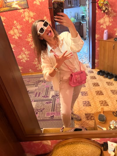 A young woman is taking a selfie in a room with floral wallpaper and wooden furniture. She is wearing a white outfit, sunglasses, and a pink purse. She is sticking her tongue out at the camera. The room appears to be a bedroom or dressing room, with a mirror reflecting the scene. The time of day is unclear, but the lighting suggests it is daytime. The location is Krasnoe-na-Volge, Russia. The woman appears to be happy and playful. There is no visible text in the image.