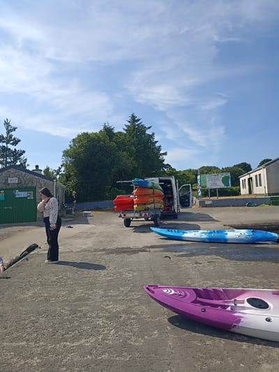 This image depicts a watersports rental location in Loughanure, Ireland. In the foreground, a pink and purple kayak rests on a concrete surface. Behind it, a blue and white kayak lies on the same surface. To the left, a young woman wearing a floral blouse and dark trousers stands facing away from the camera, holding a smartphone. Further back, a white van is parked with its side door open, loaded with several colorful kayaks stacked on a trailer. Adjacent to the van, a white building with a green door is visible. In the background, a large sign reads "LOUGHANURE ANGLERS". Trees and a clear blue sky with scattered clouds complete the scene. The environment appears to be an outdoor, possibly recreational, area near a body of water.