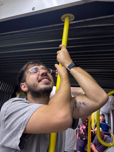 A young adult male, with a beard and eyeglasses, wears a grey t-shirt and holds a bright yellow vertical pole with both hands. He exhibits a broad smile with visible upper teeth, and his eyes appear closed or squinting as he looks upwards and to his right. A black smartwatch is present on his left wrist, and a black mountain range tattoo is visible on his upper left arm. The immediate environment is the interior of a public transport vehicle, characterized by dark, horizontally ribbed panels on the walls/ceiling and lighter panels above. In the background, other passengers are partially visible, including a male figure in a dark shirt looking towards the camera, and additional yellow poles and blue seating elements. This scene is set within a public transport vehicle at Ben-Gurion Airport, Israel.