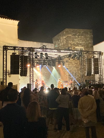 The image shows an outdoor concert at night in Loulé, Portugal. A band is performing on a stage set up with lighting trusses, speakers, and musical instruments. Several musicians are visible on stage, including a person playing a double bass, a singer at a microphone, and another musician near a keyboard. The stage is illuminated by spotlights casting colored light beams. In front of the stage, a crowd of people is gathered, watching the performance. The background consists of buildings with stone and white facades. The overall scene depicts a lively evening event in the city.