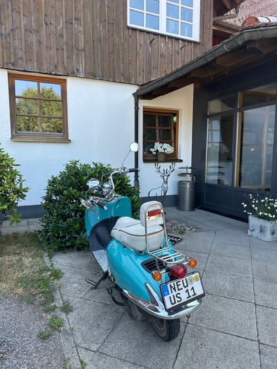 In St. Märgen, Germany, a turquoise vintage scooter is parked on a paved patio in front of a building with a wooden upper facade and a white stucco lower facade. The scooter features a white seat and a chrome rack on the back. A license plate displaying "D NEU US 11" is attached to the rear of the scooter. To the left of the scooter, a dense green bush is visible, and further left, a window with a wooden frame reflects trees and the sky. To the right of the scooter, a large windowed door with dark framing leads into the building. A potted plant with white flowers sits to the right of the door. A stylized drawing of a deer is visible on the white wall to the left of the door. The environment appears to be an outdoor area adjacent to a residential or commercial building, under natural daylight.