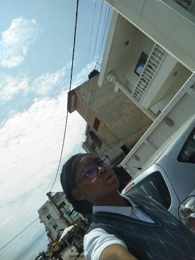 The image displays a street scene in Cotonou, Benin, captured from a low-angle perspective, tilted slightly. In the immediate foreground, a young woman of African descent is visible from the chest up, engaged in taking a selfie. She wears a dark headband, circular glasses with purple-tinted lenses, a white collared shirt, and a dark grey V-neck sweater vest with a lighter patterned design. A silver stud earring is present in her left ear. Her gaze is directed slightly upwards and to the right, with her lips parted.

Behind her, to the right, a silver or light grey passenger vehicle, possibly a hatchback, is partially visible, appearing to be parked. Directly behind the vehicle and extending upwards on the right side of the frame is a multi-story building with a light-colored, possibly stucco, facade. This building features a balcony with white balustrades and a prominent round column. Further into the background and slightly to the left, another building with a discolored, aged facade is visible, topped by a dark water tank.

The left side of the image reveals a street extending into the distance, lined with several other multi-story buildings of varying designs. A utility pole is present among these structures. Fine black electrical or communication wires crisscross the sky, running both vertically and diagonally across the frame. The sky occupies a significant portion of the upper frame, displaying a bright blue hue with scattered white cumulus clouds. The scene is brightly lit, indicating daytime conditions.