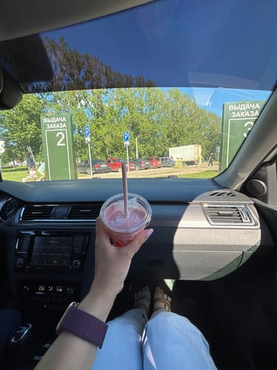 A first-person perspective shot from inside a car in Fedorino, Russia, shows a hand holding a pink smoothie with a straw. The car's dashboard is visible, with a car radio displaying "MARUSYA" and climate control settings. Outside the car, through the windshield and side windows, green trees and a bright blue sky are seen. Two green signs with white text and numbers are prominent: one reads "ВЫДАЧА ЗАКАЗА 2" (Order Pickup 2) and another, further to the right, reads "ВЫДАЧА ЗАКАЗА 3" (Order Pickup 3). Parking signs are also visible. In the background, several cars are parked, and people are walking, including one person carrying shopping bags and another with a yellow electric scooter. The overall environment suggests a sunny day in a public area, likely a parking lot or service point.