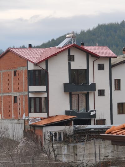 This image depicts a residential scene in the city of Ribnovo, Bulgaria, characterized by multiple buildings under an overcast sky. The central focus is a multi-story house with a white facade and dark gray trim, featuring a red tiled roof topped with a functional solar water heating system consisting of a horizontal cylindrical tank and a tilted solar panel array. This house exhibits multiple dark-framed windows of varying sizes and two prominent, dark gray rectangular balconies, one positioned above the other, with the lower balcony showing a broken or unfinished railing. To the immediate left, an unfinished brick structure is visible, exposing red bricks and partial framing, also topped with a matching red tiled roof. A small white satellite dish is attached to a lower section of the wall between these two buildings.
Further to the right, a portion of another white house with dark trim and brown-framed windows is partially visible, featuring a greenish-gray tiled roof. In the foreground, below the main structures, there is a smaller, light gray building with a steeply pitched orange tiled roof and a single window, positioned in front of a concrete retaining wall. Adjacent to this small building, piles of wood and miscellaneous debris are visible behind a lower concrete wall. Bare tree branches, indicative of a dormant season, extend into the lower foreground, with faint horizontal lines, possibly wires, crossing the very bottom of the frame. The background is dominated by a dense, dark green coniferous forest covering a gentle slope under a hazy, cloudy sky. No individuals or specific actions are observable within the frame.