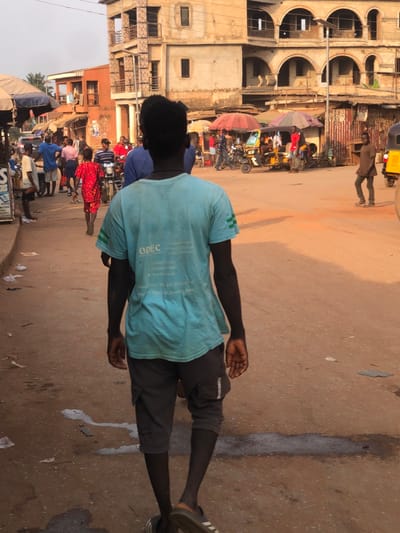 This image depicts a street scene in the city of Neni, Nigeria. In the immediate foreground, viewed from behind, is a dark-skinned individual walking away from the camera towards the right. This person wears a light blue t-shirt with "OPEC" and additional white text and numbers visible on the back, dark grey cargo shorts, and dark slides.
The ground is an unpaved, reddish-brown surface, showing scattered pieces of litter and several dark, wet stains.
To the left, a collection of individuals is present, including a person in a blue shirt and a woman in a red patterned dress walking away. Market stalls with light-colored and striped awnings line the street, with a partially visible sign. A motorcycle is positioned among the pedestrians. Further down this left side, a multi-story building with a reddish-brown exterior is visible.
On the right side of the street, more individuals are observed, along with at least two yellow auto-rickshaws (keke-napeps) and other motorcycles. Market stalls are present under red and striped umbrellas. An individual in a brown shirt walks towards the right.
In the background, a prominent multi-story building with a light beige or off-white facade features multiple arched openings on its upper levels. The sky is clear, and the scene is illuminated by natural daylight.