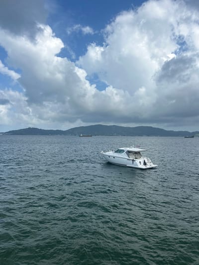 A white motor yacht is moored in dark, rippling water. The yacht has a hardtop and appears to be unoccupied. In the background, a lush green, mountainous coastline stretches across the horizon under a cloudy sky. Two cargo ships are visible in the distance, one to the left and one to the right of the yacht. The water is choppy, indicating a moderate breeze. The scene is a wide shot, capturing the expanse of the sea and the distant land. The location is the sea in the vicinity of Ban Hin Chang, Thailand.