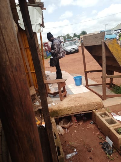 In Agwala, Nigeria, a person stands next to a wooden structure, looking down at their phone. They are wearing a patterned jacket and a black head covering. The ground is a mix of red dirt and cracked pavement. A small wooden stool sits to the left of the person. In the background, several cars are parked on a dirt road, including a light blue vehicle and a silver car. A bright blue bucket is visible on the pavement near the wooden structure. The scene appears to be outdoors, possibly a marketplace or roadside stall, given the presence of vehicles and the casual setup. The sky is partly cloudy, suggesting it is daytime. There is some debris and trash scattered on the ground in the foreground, with a visible concrete block structure. A sign with text, partially obscured, is visible on a building in the distance.