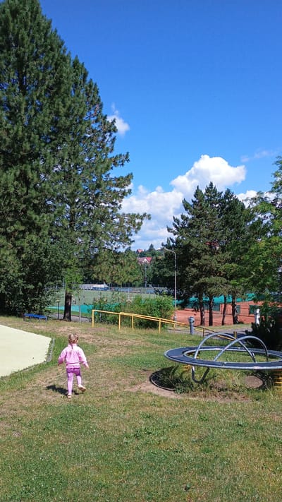 A young child with blonde hair in a braid, wearing a pink tracksuit, walks away from the camera across a grassy area in what appears to be a park or playground. The child is moving towards a black and silver merry-go-round. In the background, there are trees, a fence, a sports field, tennis courts with red clay surfaces, and a few houses with red roofs on a distant hill. The sky is bright blue with scattered white clouds. The scene is illuminated by daylight, suggesting it is a clear, sunny day. The location is the Sokolov Municipal Stadium sports complex in Sokolov, Czechia, specifically the area between the stadium and the tennis courts.