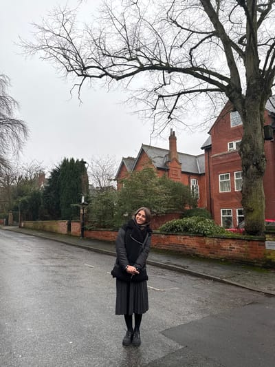 A woman stands on a wet street, dressed in a black jacket and pleated skirt, smiling towards the camera. Behind her, a red brick building with white-framed windows and a steeply pitched roof rises amidst green bushes and bare trees. A tall, leafless tree dominates the left side of the frame. The sky is overcast, giving the scene a muted, cool tone. The pavement is slick, reflecting the somber light.