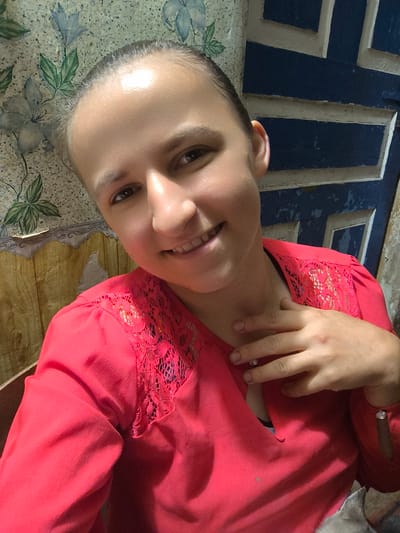 The media file is a portrait of a young woman indoors, with her head and shoulders filling most of the frame. She has her hair pulled back, and her eyes are looking directly at the camera with a gentle smile. She is wearing a bright red blouse with lace detailing on the shoulders and sleeves, and a diamond-like embellishment on her wrist. Her left hand is resting on her collarbone, and her right hand is placed on her chest. The background is a mix of textures and colors. To the left, there is a floral wallpaper with blue and green patterns, and to the right, a blue and white patterned surface that appears to be a door or wall paneling. The lighting suggests an indoor setting, possibly during the daytime. The woman's expression conveys a sense of warmth and openness. The overall impression is that of a casual, personal photograph, likely a selfie. There is no visible text in the image.