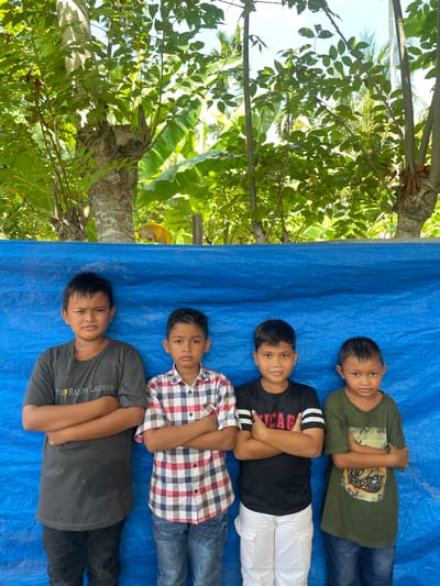 Four young boys stand with their arms crossed in front of a vibrant blue tarp. The boy on the far left wears a dark grey t-shirt with the text "POLO RALPH LAUREN" visible, and dark pants. The second boy from the left is dressed in a red, white, and blue plaid shirt with dark pants. The third boy wears a black t-shirt with white stripes on the sleeves and "CHICAGO" in red lettering, paired with white cargo pants. The boy on the far right is in a green t-shirt with a graphic design and blue jeans. They are positioned outdoors with lush green foliage and tree trunks visible in the background, suggesting a tropical environment. In the distance, a yellow object is partially visible. The image appears to be taken in Peudaya, Indonesia, judging by the tropical flora and the style of the children's attire.