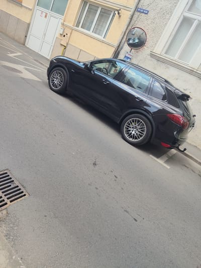 A black Porsche Cayenne is parked on an asphalt street in Arad, Romania. The car is angled towards the right of the frame, with its rear visible. It has silver multi-spoke wheels with yellow brake calipers. A trailer hitch is attached to the rear bumper. The street has a metal drain grate in the lower left corner. The building on the right is light-colored with white-trimmed windows. A street sign with "STR. VASILE GOLDIS" is visible on the wall, along with a convex mirror reflecting the street and another black car. The building on the left is yellow with white doors and windows. The scene appears to be during the daytime, with natural light casting shadows. The weather seems clear. No people are visible.