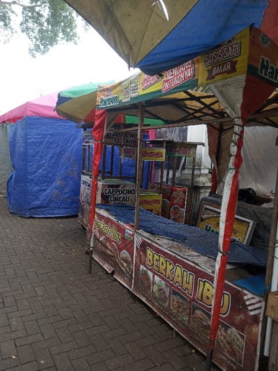 The image depicts an outdoor vendor area in the city of Cianjur, Indonesia. In the foreground, a prominent food stall is visible, characterized by a multi-colored canopy in shades of faded yellow, blue, red, and white. The canopy displays branding, including "Harmini," "SOSIS SAPI BAKAR" (Grilled Beef Sausage), and "NENDANG UNTUNGNYA!" (Kick-in the profits!). The stall's counter is covered by a red and white banner featuring "BERKAH IBU" (Mother's Blessing) alongside images of various food items such as burgers, fried chicken, noodles, and hotdogs. Inside the stall, shelves hold signs indicating offerings like "CAPPUCINO CINCAU" (Cappuccino Grass Jelly) and other Indonesian food names. A blue tarp covers a section of the counter, suggesting the stall is either closed or items are protected. To the left, a second vendor stall, covered in a large blue tarp and topped with a pink/red canopy, is partially visible. The ground is paved with dark grey interlocking bricks. The sky is overcast, and green foliage from trees is visible in the upper left corner. No individuals are present in the scene.