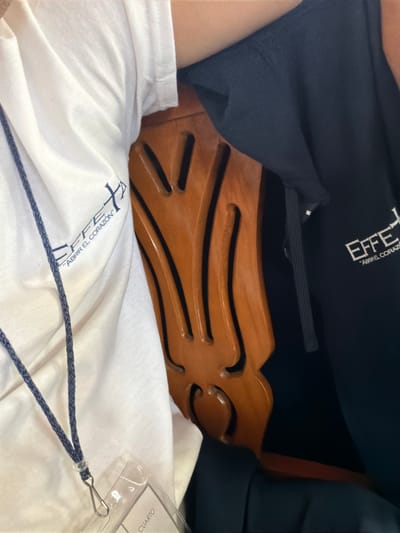The image shows a close-up of clothing items and a wooden chair back. A white t-shirt with a blue logo is visible in the foreground, along with a lanyard and a clear plastic ID badge. A black sweatshirt with white lettering rests behind the chair back. The chair back is made of brown wood and has an intricate design with several horizontal slots. The exact location within Santa María Zacatepec, Mexico, cannot be determined from the visual information provided.