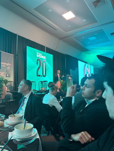 Attendees seated at a table with soup bowls and silverware are observing a presentation at the "Project Inspire 2.0 Convention." A large screen displays the event logo and branding. Some individuals are using mobile devices. The setting appears to be a conference hall or ballroom with ceiling lights and dark drapery. The perspective is from a slightly elevated angle, showing a partial view of the attendees' heads and shoulders.