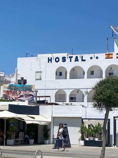A clear, sunny day in Conil de la Frontera, Spain, showcases a white building with arched balconies and dark blue lettering spelling "HOSTAL" above the second-floor windows. Two star-shaped decorations are also present on the building. A Spanish flag and a green flag are mounted on the right side of the building. To the left, a sign for "Restaurante PLAYA" displays images of fish. A tree with dense green foliage grows in the foreground, partially obscuring the building. In front of the building, a person walks with a rolling suitcase and a backpack. A white awning covers an outdoor seating area to the left, where several people are seated at tables. A metal rack is visible in the lower left corner.