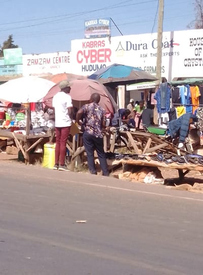 The image depicts a bustling roadside market scene under a clear blue sky in South Kabras ward, Kenya. In the foreground, a gray asphalt road extends across the bottom, with a small, light-colored piece of debris lying on its surface.
Immediately adjacent to the road on the right, several makeshift market stalls are constructed from rough wooden poles and planks. These stalls are covered by various large umbrellas or tarpaulins providing shade: a white one on the far left, a brown one in the center, and a blue one further right.
Multiple individuals are present among the stalls. Two men are prominent in the mid-ground: one male, facing right, wears a white t-shirt, maroon trousers, and a light-colored cap. The second male, positioned slightly to his right and facing away, wears a patterned short-sleeve shirt and dark trousers, with hands near his hips. Other less distinct individuals are visible deeper within the market area, engaging in activities such as arranging goods or interacting with others.
The market displays a variety of merchandise. On the left, household items, possibly kitchenware or plastic containers, are arranged on a stall. A yellow sack is visible beneath this stall. On the right, clothing items including t-shirts, jeans, and a blue puffer jacket are hung on racks and displayed on tables. Numerous pairs of shoes or sandals are laid out in rows on a low wooden platform.
In the background, behind the market stalls, a line of white commercial buildings is visible. Prominent signage on these buildings includes "KABRAS CYBER HUDUMO," "ZAMZAM CENTRE," "Dura Coat ASK THE EXPERTS," and "KAKUNGA FARM." A smaller sign for "KABRAS CYBER COMPUTER TRAINING CENTRE" is mounted above the main building line. A wooden utility pole stands vertically on the far right, with power lines visible across the top of the frame.