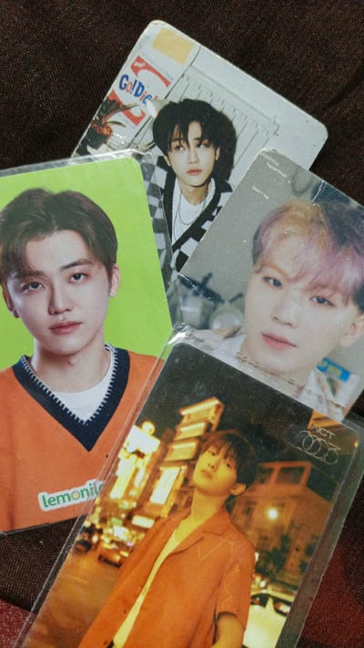 The image shows a close-up, overhead view of several K-pop photocards layered on top of each other. The background appears to be a dark, textured fabric, possibly a blanket or tablecloth.
From top to bottom, the visible photocards are:
1. A card with the text "Go!Die" in red lettering. The image features a young man with dark hair, wearing a black and white checkered jacket over a white t-shirt and a silver chain necklace.
2. A card with a bright green background, featuring a young man with brown hair wearing an orange v-neck sweater over a white t-shirt. Text in white on the bottom left reads "lemoni".
3. A card with a greyish background, partially obscured by the other cards. Text at the top reads "EVENTEEN" and "Digital Single". An image of a person with light pink hair is visible.
4. A card with a dark background and a cityscape at night, illuminated by warm lights. The image shows a young man with dark hair wearing an orange short-sleeved button-up shirt over a white t-shirt. The text "NCT" and "2023" are visible in the top right corner of this card.
The image does not contain any information or visual cues that indicate the location is Palohlada, Indonesia.