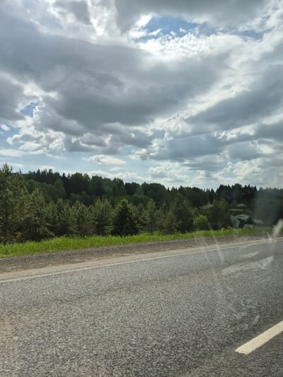 The image shows a view from a car on a road, likely in a rural setting. The road is made of asphalt and has a white line marking the edge. Next to the road is green vegetation. On the other side of the vegetation is a dense line of trees. Above the trees is a sky filled with white and gray clouds, with patches of blue sky visible. The photo appears to have been taken through the car window and is slightly tilted. There is a reflection in the car window in the top right corner. There is no indication of the location being Chernovskoye, Russia.