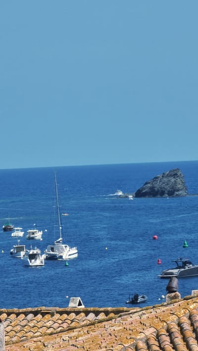 This media file depicts a serene seascape with a clear blue sky and calm blue waters. Several boats are anchored in the bay, including a white catamaran with a sail, and several smaller white motorboats. A rocky outcrop juts out from the sea on the left side of the frame, with a small white structure visible near its base. In the foreground, a tiled roof extends into the frame from the right, suggesting the vantage point is from a building overlooking the bay. The scene appears tranquil, with no clear indication of human activity beyond the presence of the boats. The lighting suggests a sunny day, likely midday given the brightness and lack of shadows. The location is identified as Cadaqués, Spain, a coastal town known for its picturesque bays and historic architecture. There is no visible text in the image.