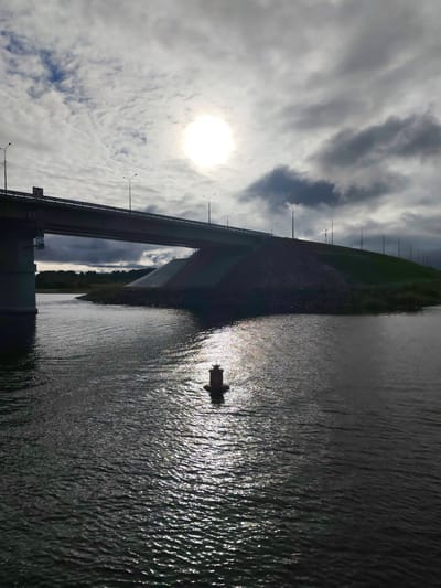 A view of a bridge in Нифантово, Russia. The image is taken from the water, showing a waterway in the foreground with a single red buoy floating on the surface. Sunlight reflects off the water. A bridge spans the waterway, supported by concrete pillars and flanked by a green embankment. Streetlights line the top of the embankment. The sky is overcast with the sun shining through the clouds.