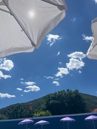 This is a low-angle, outdoor photograph taken in Varvara, Bulgaria, on a sunny day. The image is dominated by a bright blue sky filled with scattered white clouds. Two large white umbrellas are visible, one in the upper left and another partially visible in the upper right, obscuring parts of the sky. Sunlight shines through the fabric of the umbrella on the left, creating a lens flare effect. In the middle ground, a lush green, tree-covered hillside rises, with a few houses and a brick chimney peeking through the foliage. The lower portion of the image shows a dark blue surface, possibly a pool, with four open purple umbrellas lined up. These umbrellas have white branding that appears to read "Milka Ice Cream." The scene suggests a resort or poolside area on a clear, warm day.
