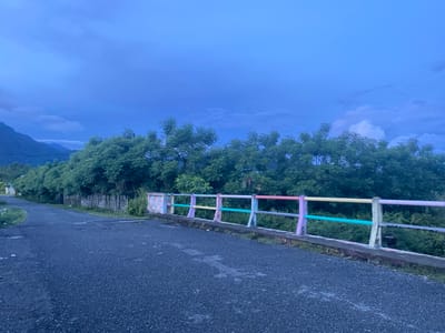 The image depicts a twilight scene on a road in Aneuk Paya, Indonesia, with a mountain range visible in the distance. A dark asphalt road curves to the left, leading into a densely vegetated area. On the right side of the road, a concrete railing separates the road from the vegetation. The railing is decorated with brightly colored horizontal bars in pastel shades of pink, yellow, green, and purple, interspersed with concrete posts. The sky above is a deep, moody blue, with hints of clouds. The overall impression is one of a rural or semi-rural setting as dusk settles.