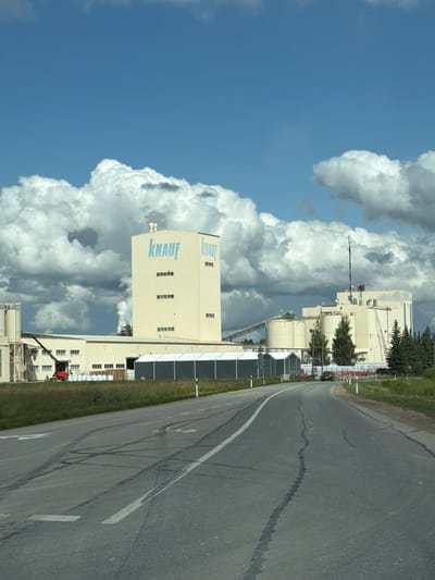 A medium-shot image captures the exterior of a large industrial complex against a backdrop of a bright blue sky dotted with dramatic, fluffy white clouds. The main structure is a tall, light-colored building with "KNAUF" prominently displayed in light blue lettering. Smaller, elongated windows are visible in rows on its facade. To the left and right of this central building are lower, interconnected industrial structures with varying shapes and sizes, featuring pipes, storage tanks, and conveyors. A red crane is positioned near the left side of the complex, and several white, bagged materials are stacked on the ground in front of a long, tent-like structure. The foreground is dominated by a two-lane asphalt road with visible tire marks and lane markings. A few cars are seen on the road in the distance, approaching the industrial site. The surroundings include green grass and scattered trees, with a line of coniferous trees visible on the right side of the image. The scene appears to be a bright, possibly sunny day, given the strong lighting and shadows. The image is taken from a slightly elevated perspective, looking towards the industrial complex from the road. The location is identified as Jaunsaurieši, Latvia.