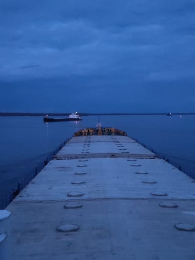 The image presents a wide, low-light view from the stern of a large vessel, likely a barge or cargo ship, on a substantial body of water near Gorushka, Russia. The foreground shows the broad, flat deck of the viewer's vessel, covered with evenly spaced, circular hatch covers running down its length. The deck surface appears light-colored, possibly due to low light or a thin layer of frost.
In the mid-ground, to the left, another large cargo ship is visible, either navigating or anchored. It has a dark hull and a lighter-colored superstructure, with several illuminated lights on its deck and mast reflecting on the calm, dark blue water. Further in the distance, a faint light source is visible on the water to the right, suggesting another distant vessel or buoy.
The water itself is a deep, tranquil blue, mirroring the color of the overcast sky above. A dark, indistinct landmass or shoreline stretches across the horizon. The sky is a uniform, dark blue, indicating either dusk or dawn, often referred to as the "blue hour," with subtle cloud formations adding texture. The artificial lights on the vessels stand out against the dim natural light. No text is visible in the image. The scene evokes a quiet, solitary atmosphere on a waterway during twilight hours.